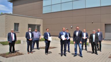 10 men in suits stand in front of a building. Two are holding a document in their hands.