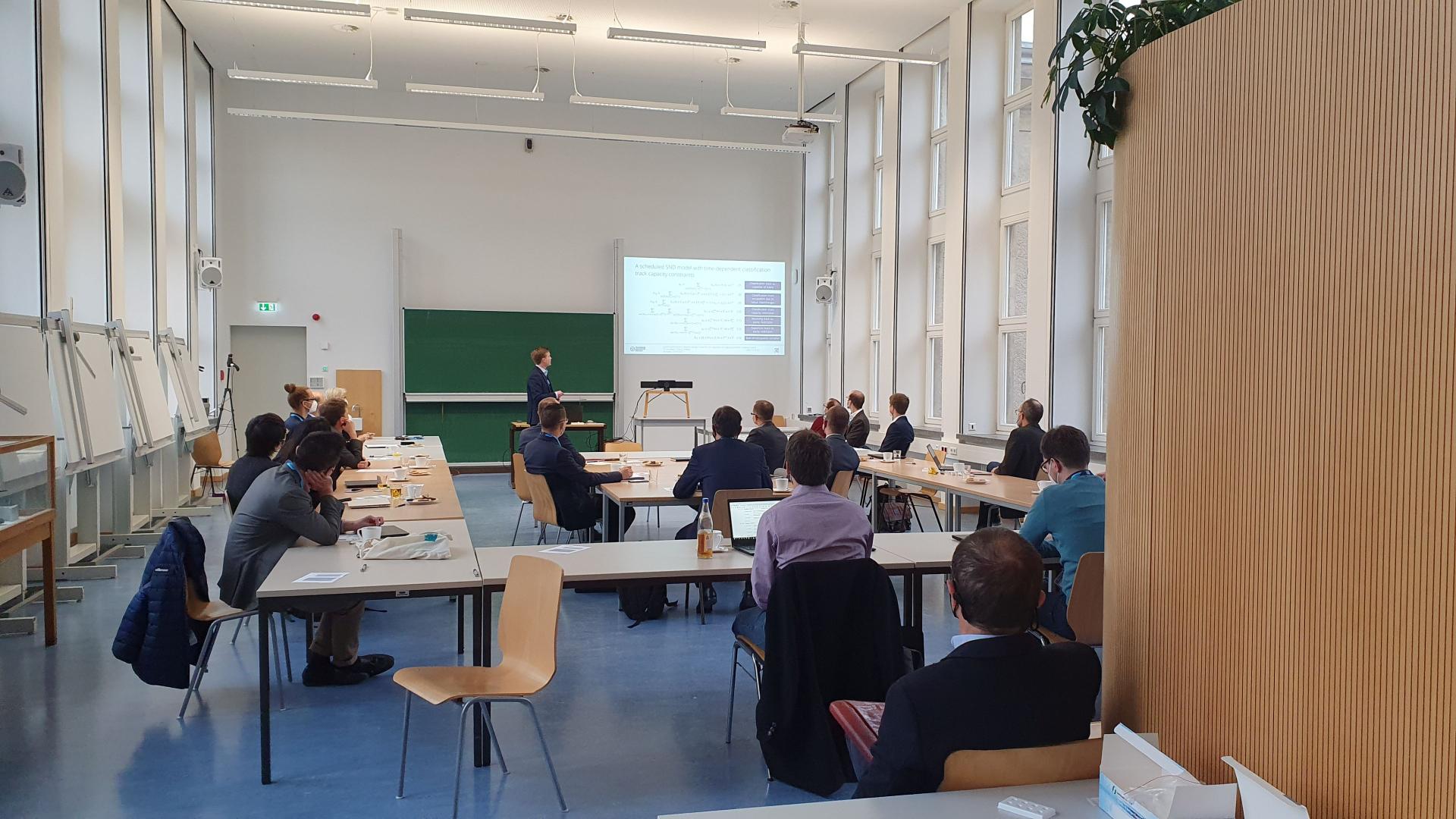 A seminar room with people. One of them is giving a lecture at the front.