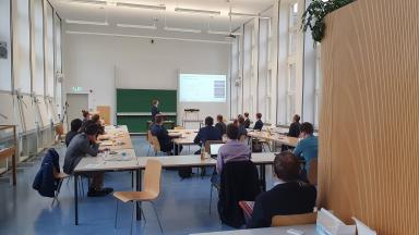A seminar room with people. One of them is giving a lecture at the front.