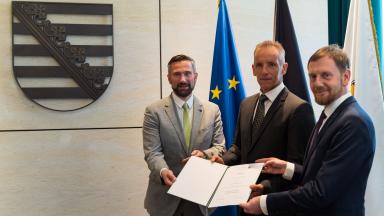 3 men stand side by side holding a certificate together. They are all wearing suits and ties.