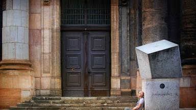 A man sits on the floor, leaning against large granite stones in front of a wooden door.