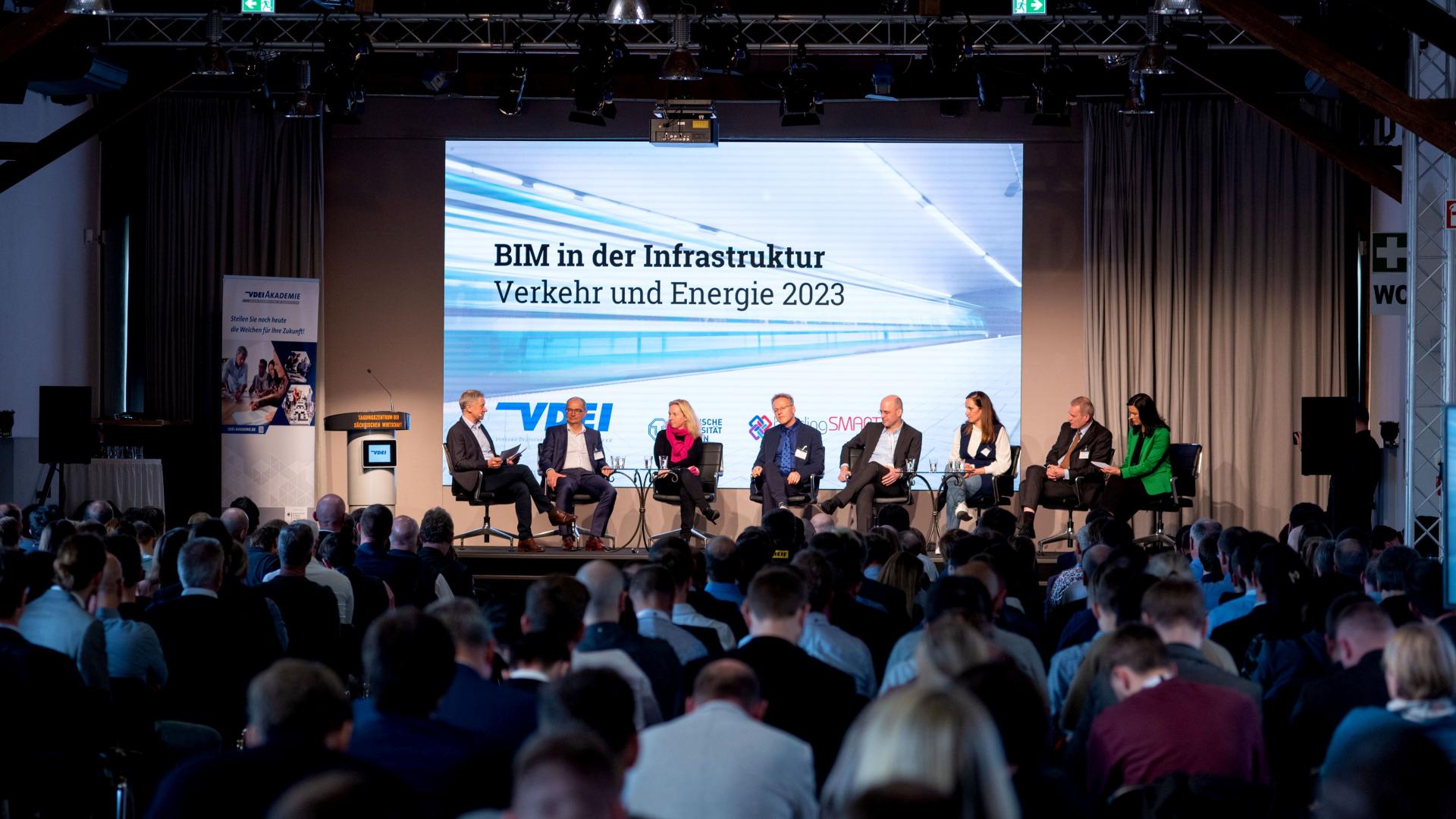 A panel discussion on a stage with 8 participants in front of many spectators
