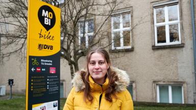 A young woman in a yellow jacket stands in front of a sign and building