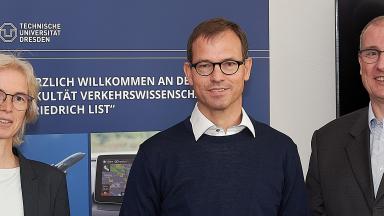 Three people stand in front of a rollup, a woman on the left. A man in the middle and on the right