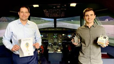  Two men stand in an aircraft cockpit. One of them holds a certificate in his hands.
