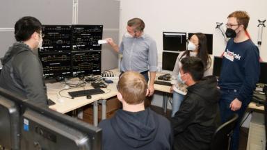  6 People stand and sit in front of a technical device.