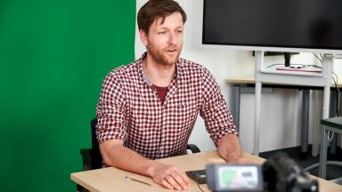 A man sits in front of a camera and a green wall.