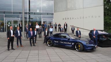A group of people stand in front of a building, along with a Porsche and a car cockpit.