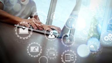 A man sits at a laptop at a table. In the foreground, symbols representing technology and study