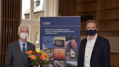 Zwei Männder mit Mundschutz nebeneinander, einer hält Blumen, in der Mitte ein Rollup