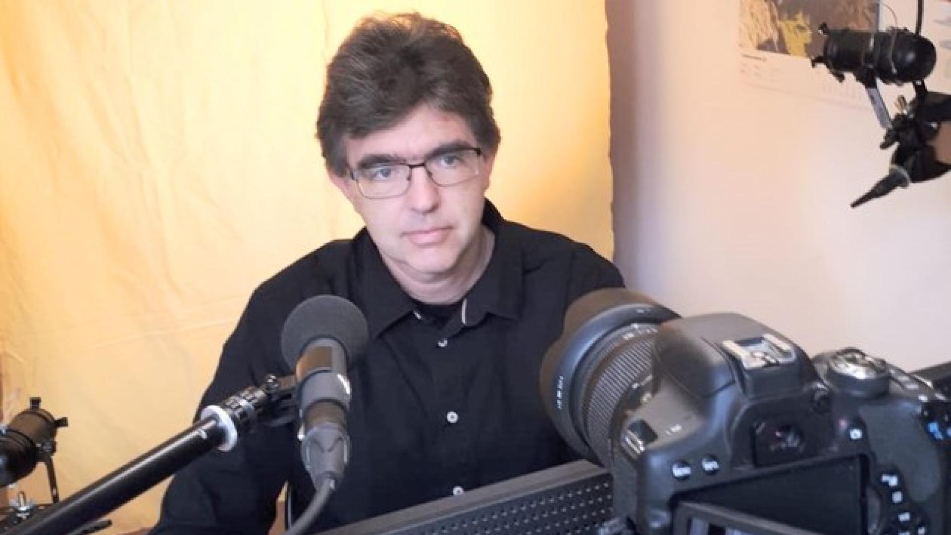 A man sits behind a mikrofon and a laptop. He is wearing glasses.