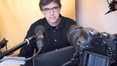 A man sits behind a mikrofon and a laptop. He is wearing glasses.