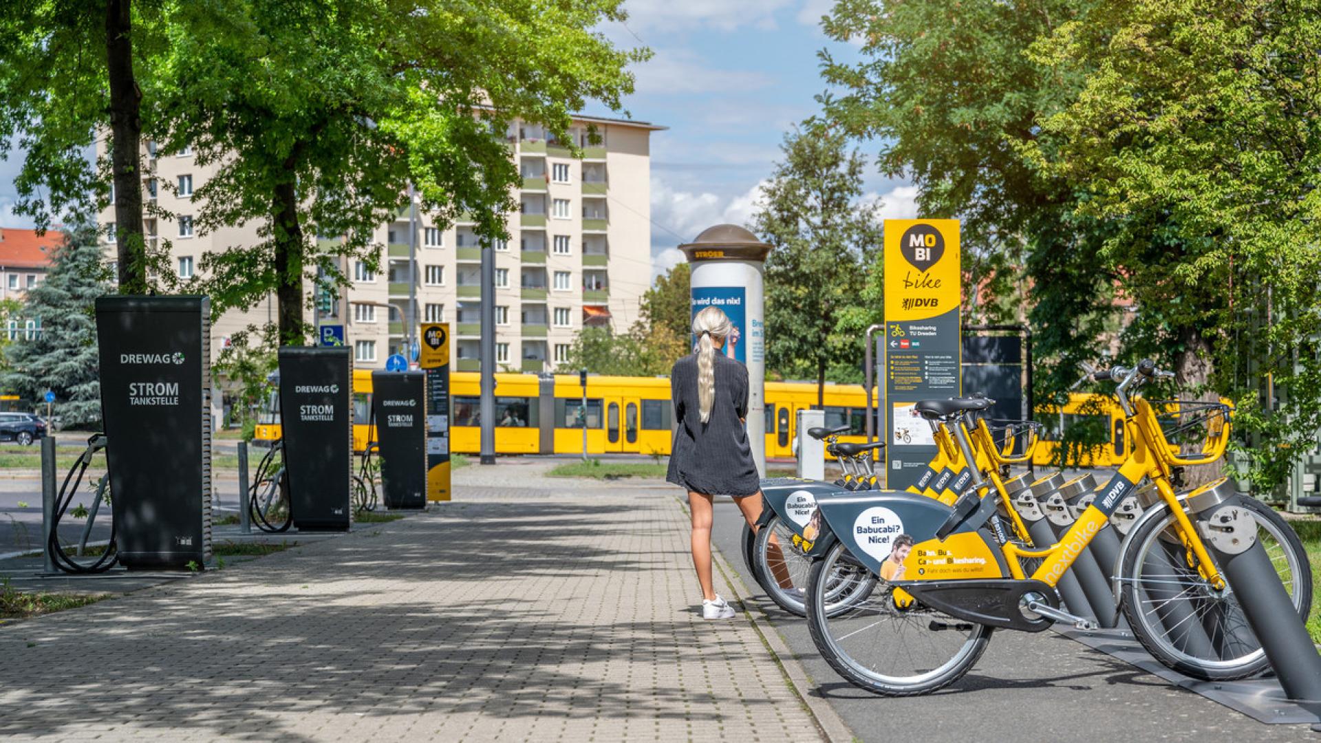 Im Hintergrund eine Tram, rechts parken E-Bikes, links sind Stromtankstellen, mittig eine Frau