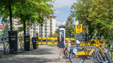 Im Hintergrund eine Tram, rechts parken E-Bikes, links sind Stromtankstellen, mittig eine Frau