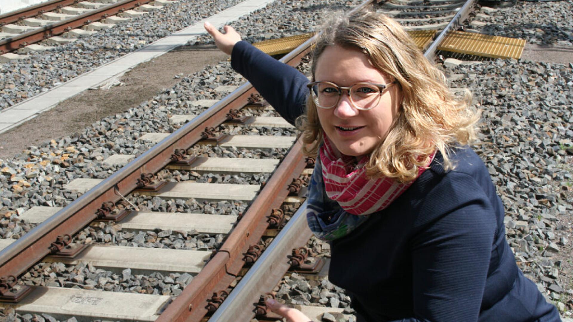 Eine Frau mit blonden Haaren und Brille hockt an einer Bahnschiene und zeigt auf etwas.