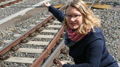 A woman with blond hair and glasses squats by a railway track and points at something.