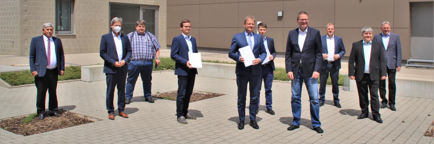 10 Männer in Anzügen stehen vor einem Gebäude. Zwei halten ein Dokument in Händen.