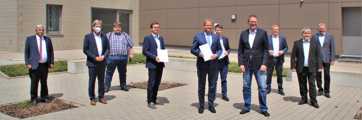 10 men in suits stand in front of a building. Two are holding a document in their hands.
