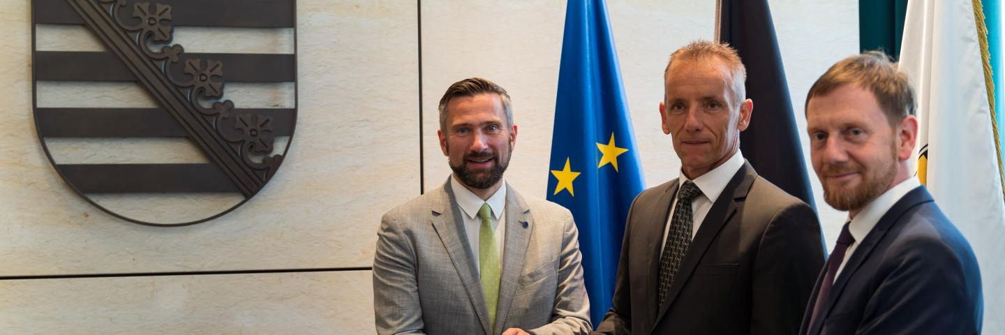 3 men stand side by side holding a certificate together. They are all wearing suits and ties.