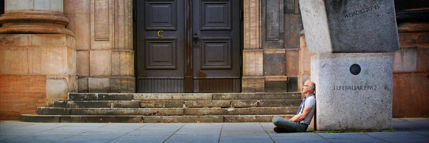 A man sits on the floor, leaning against large granite stones in front of a wooden door.