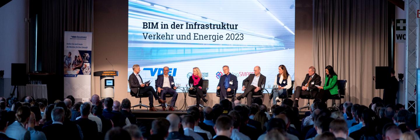 A panel discussion on a stage with 8 participants in front of many spectators