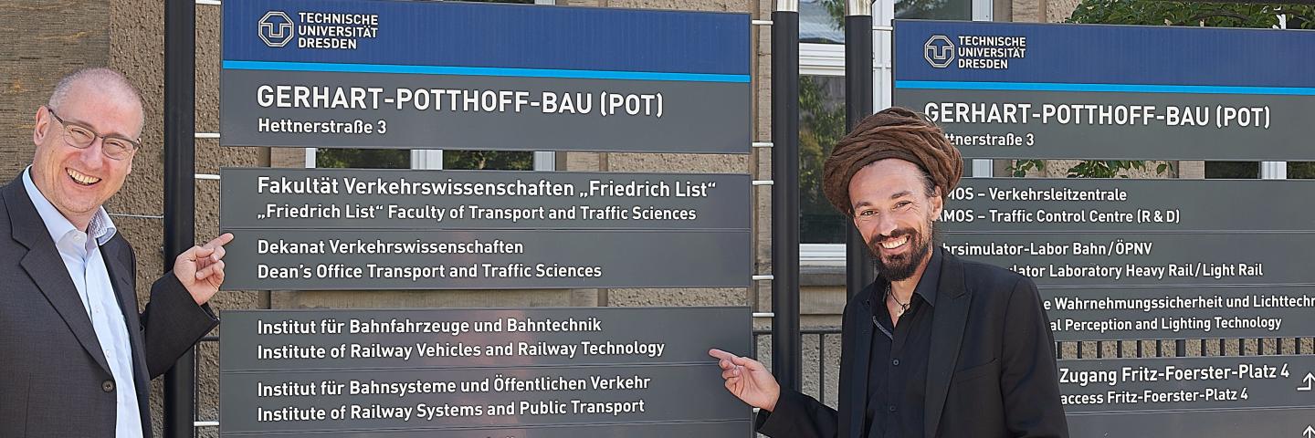 Two men stand to the left and right of a notice board.