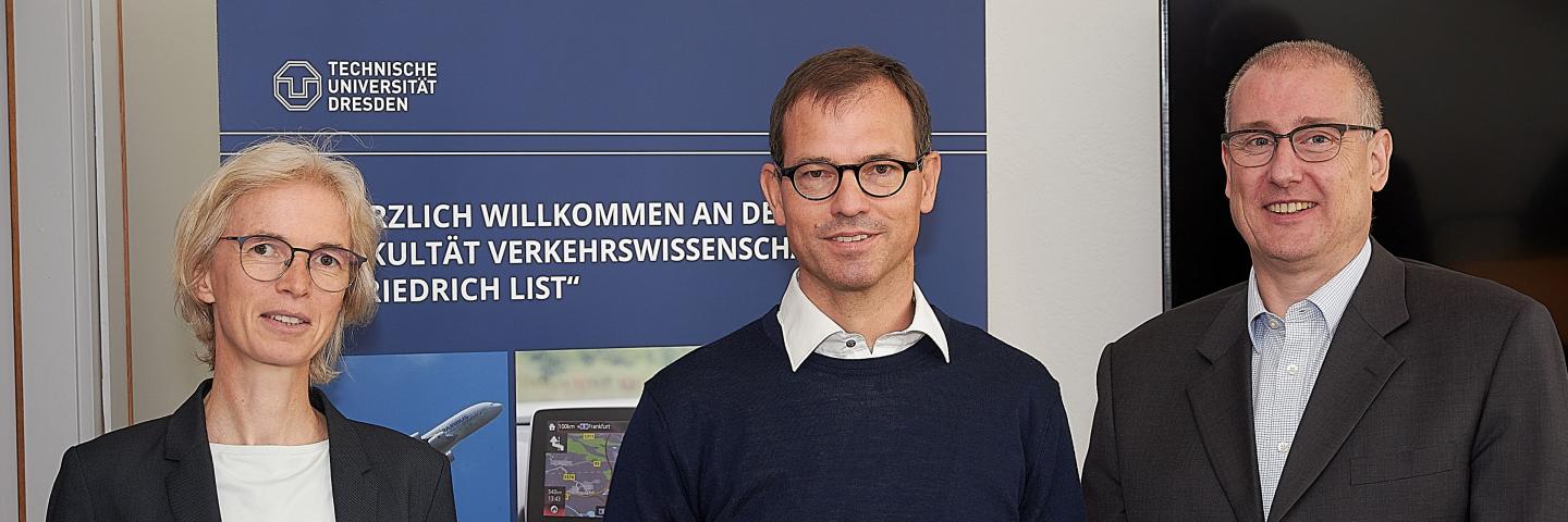 Three people stand in front of a rollup, a woman on the left. A man in the middle and on the right