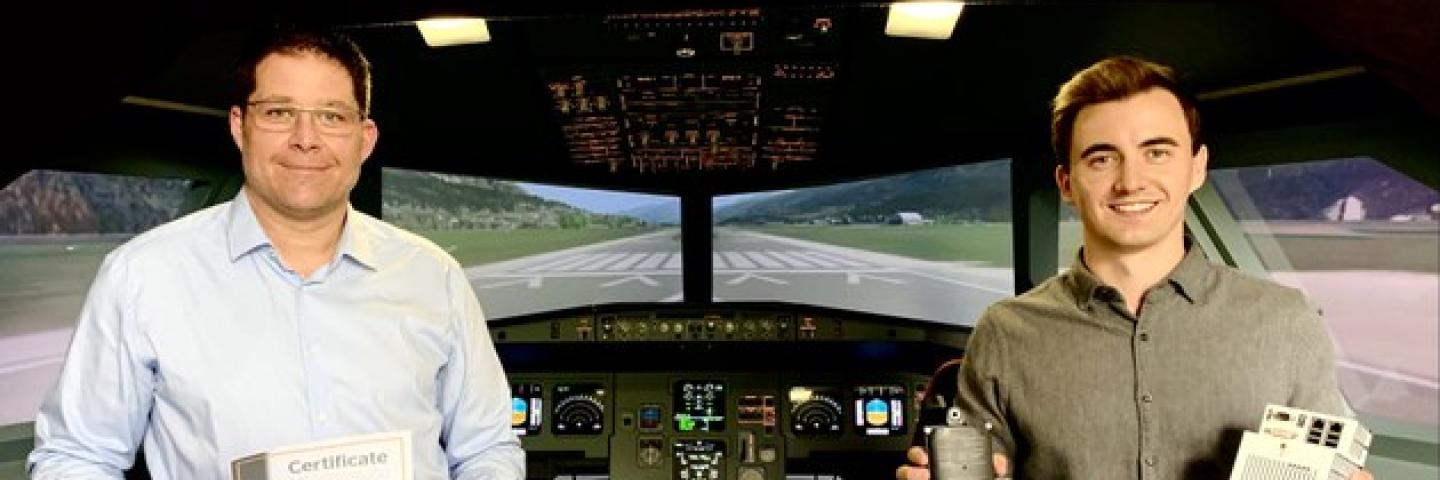  Two men stand in an aircraft cockpit. One of them holds a certificate in his hands.