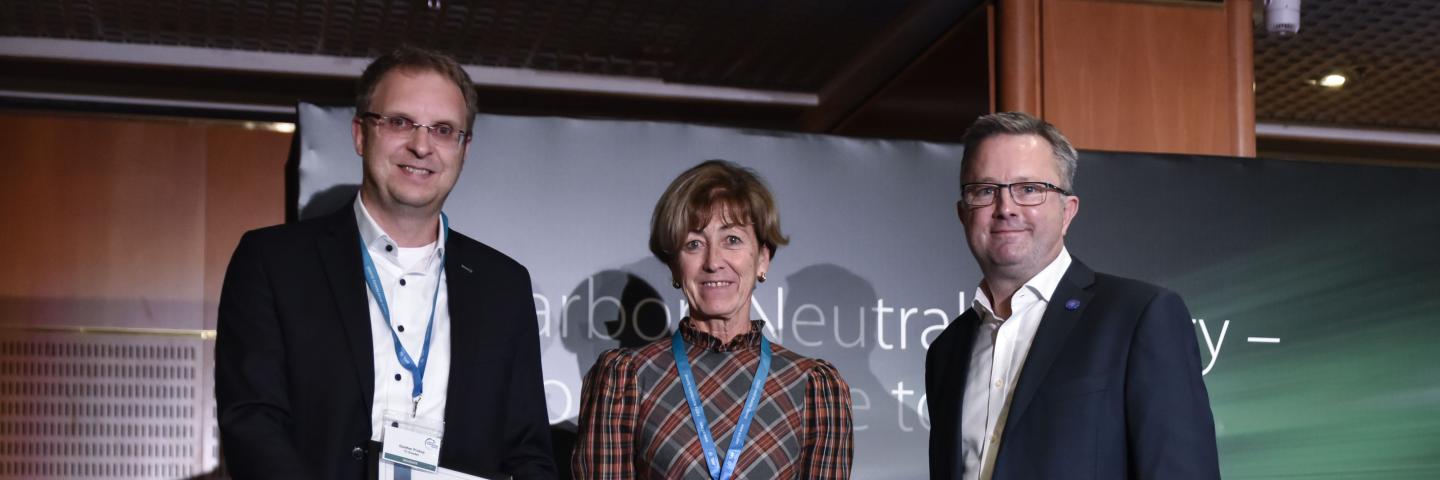 Zwei Männer und eine Frau in der Mitte stehen bei einer Preisverleihung