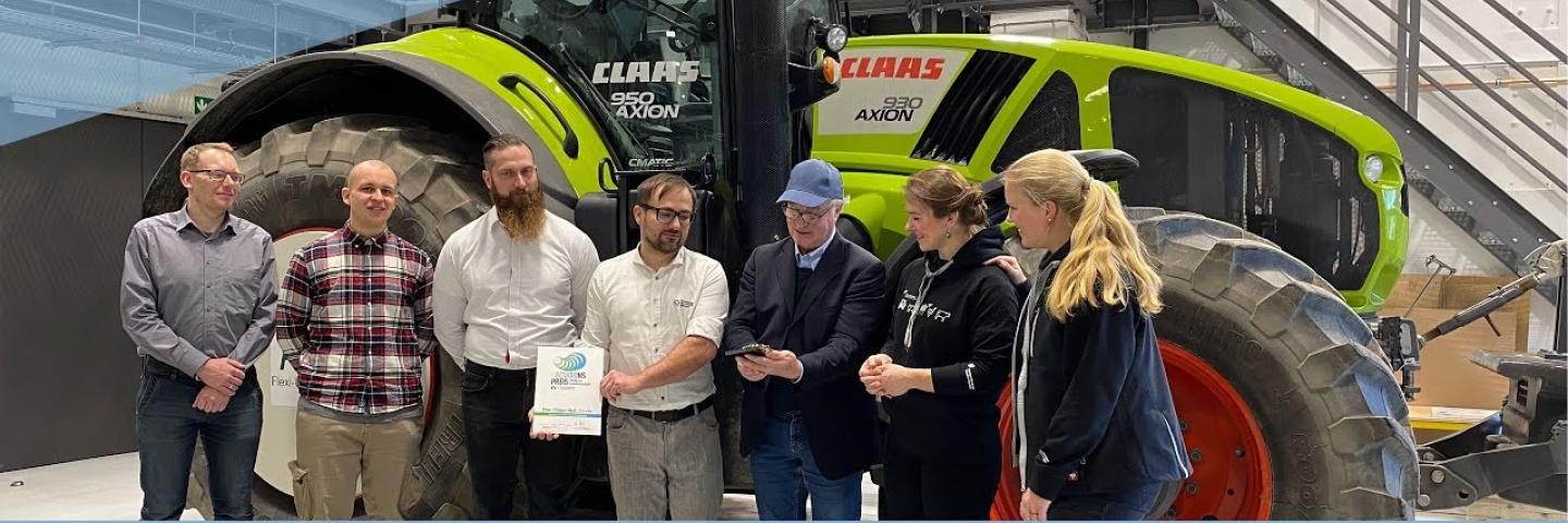 5 men and 2 women stand in front of a tractor. One man is holding a prize, another a certificate.
