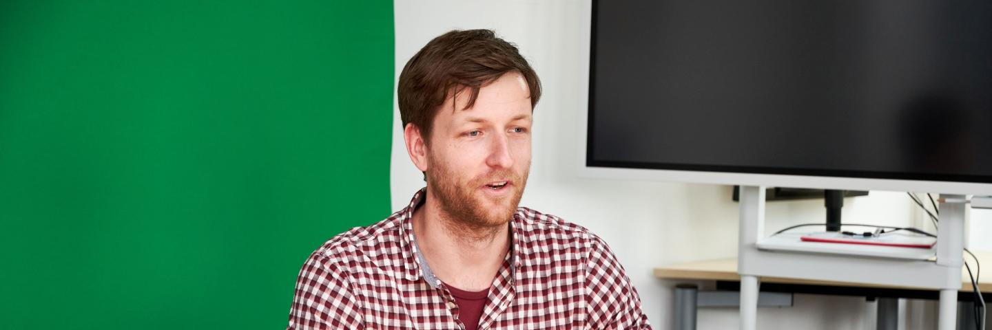 A man sits in front of a camera and a green wall.