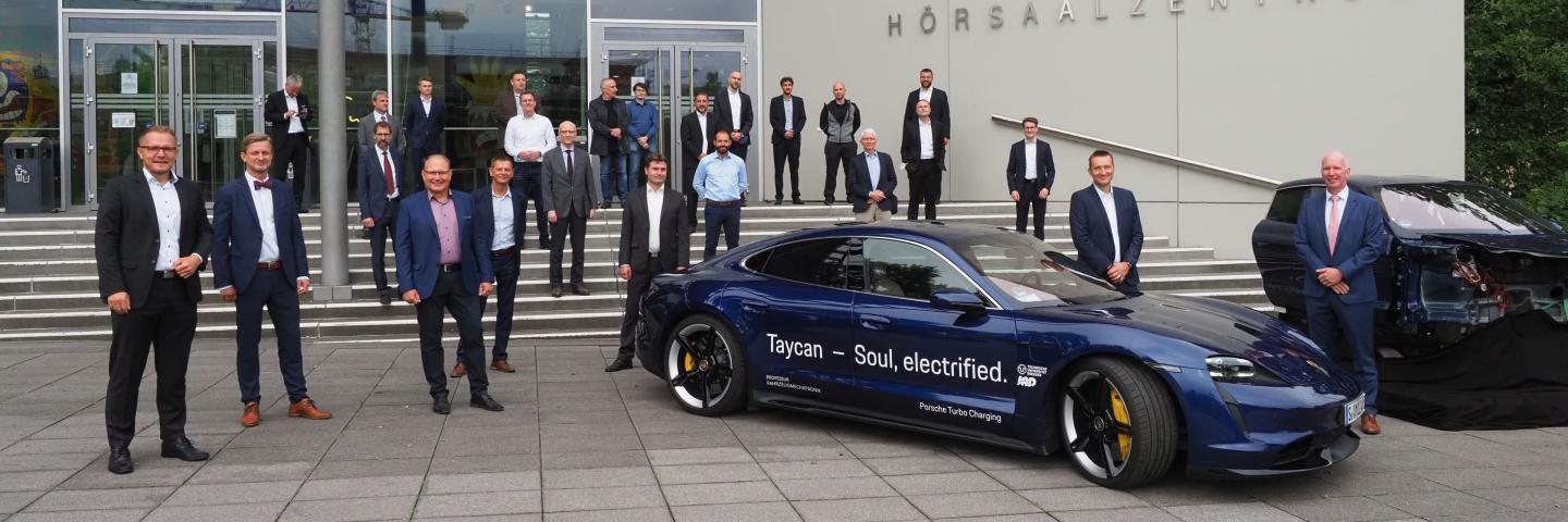 A group of people stand in front of a building, along with a Porsche and a car cockpit.