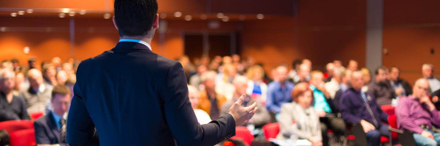 The photo shows a speaker giving a presentation to a large group.