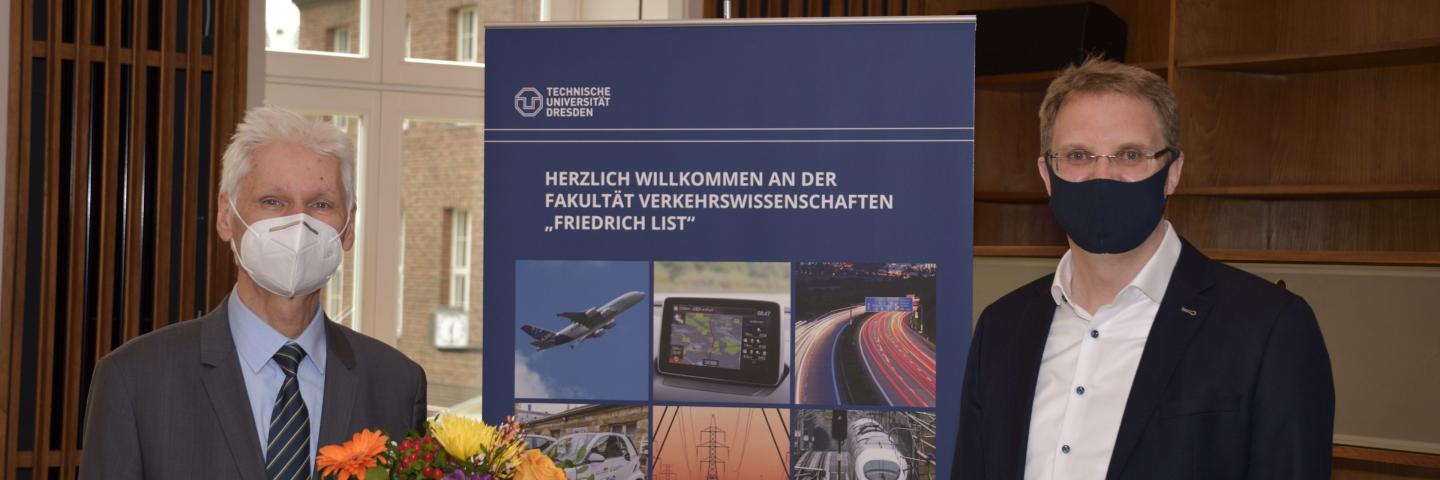 Zwei Männder mit Mundschutz nebeneinander, einer hält Blumen, in der Mitte ein Rollup