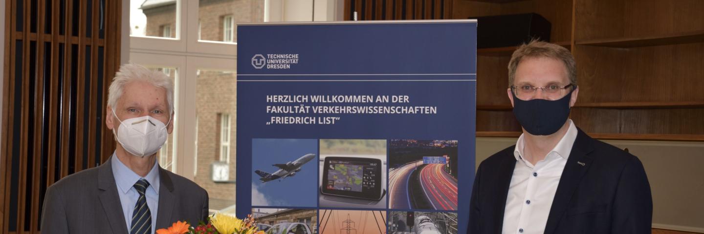 Two men with face masks next to each other, one holding flowers, a rollup in the middle.