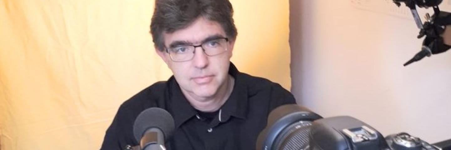 Ein Mann sitzt hinter einem Mikrofon und Laptop. Er trägt eine Brille und ein dunkles Hemd.