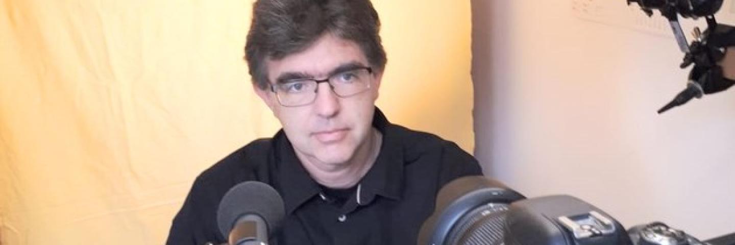 A man sits behind a mikrofon and a laptop. He is wearing glasses.