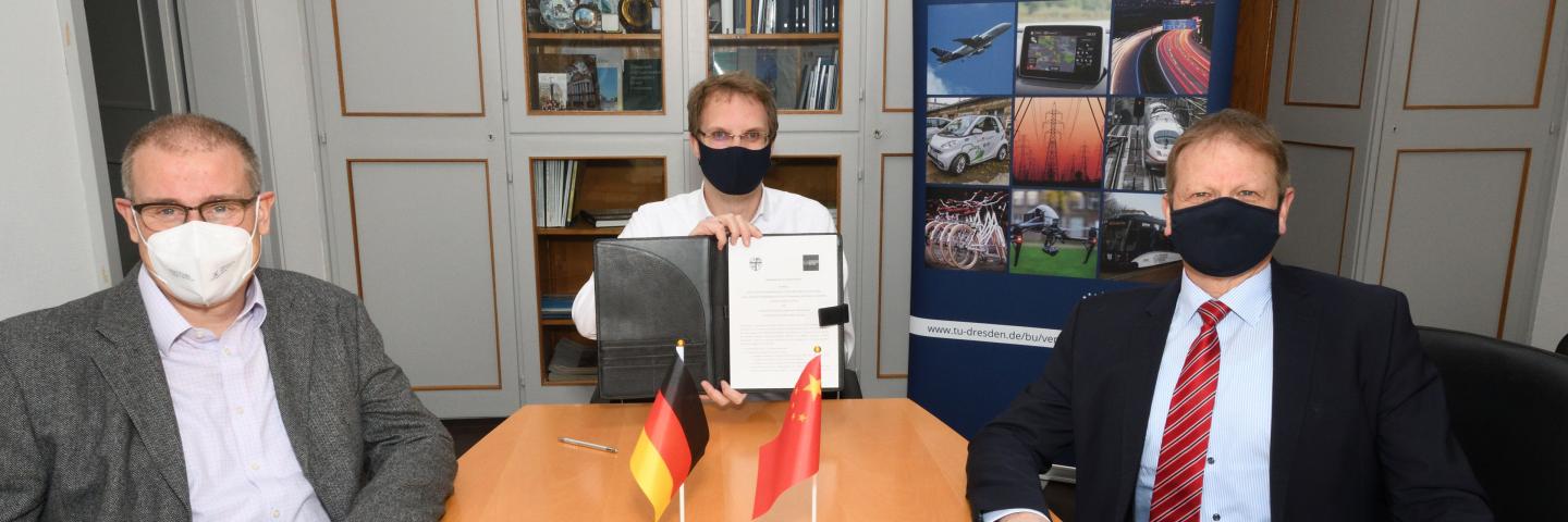 Drei Männer sitzen mit Mundschutz in einem Büro. Der mittlere hält ein Dokument hoch.