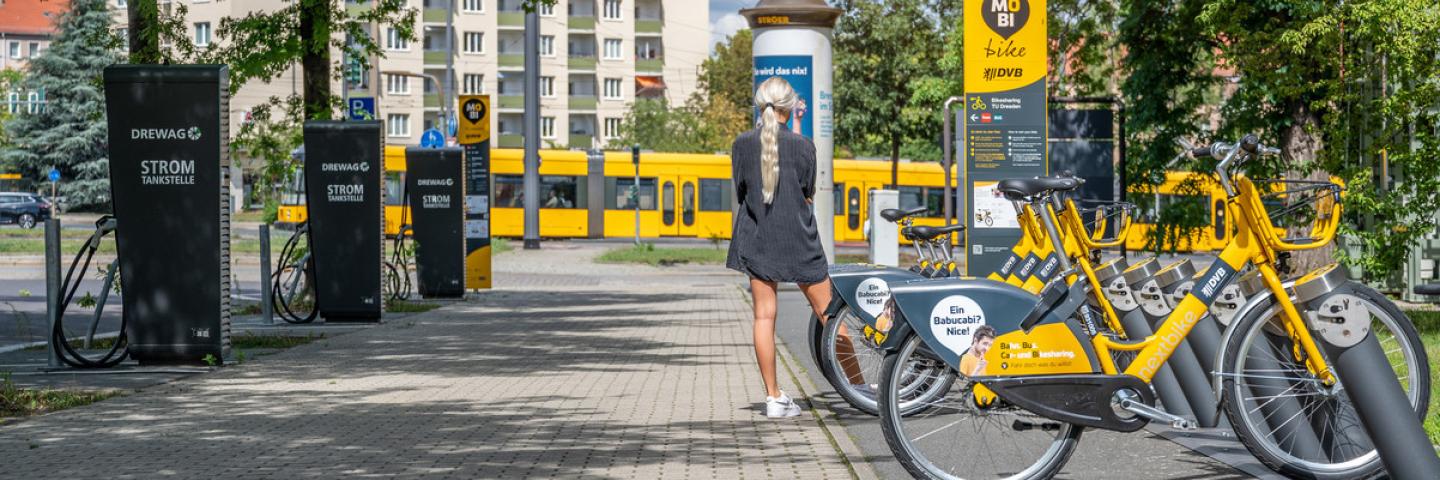 A tram in the background, e-bikes parked on the right, charging stations on the left, a woman