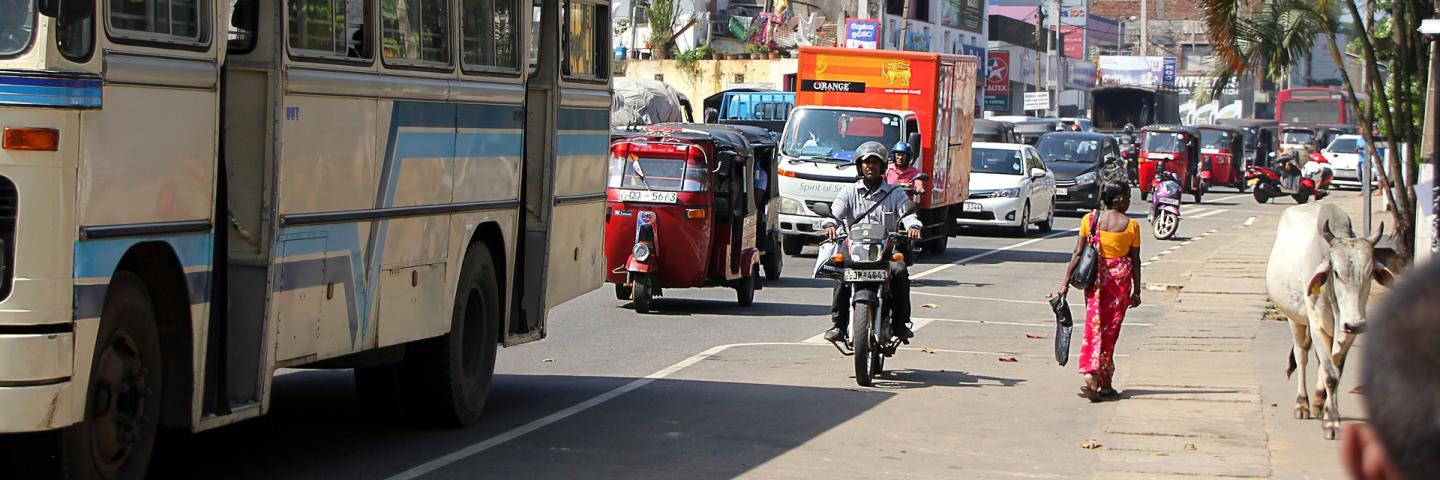Eine volle Straße in Srki lanka mit einem Bus, Autos, ein Motorrad, eine Frau und Kuh am Straßenrand