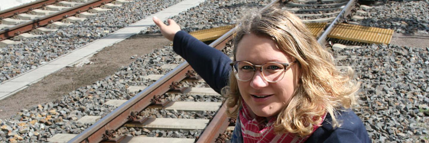 Eine Frau mit blonden Haaren und Brille hockt an einer Bahnschiene und zeigt auf etwas.